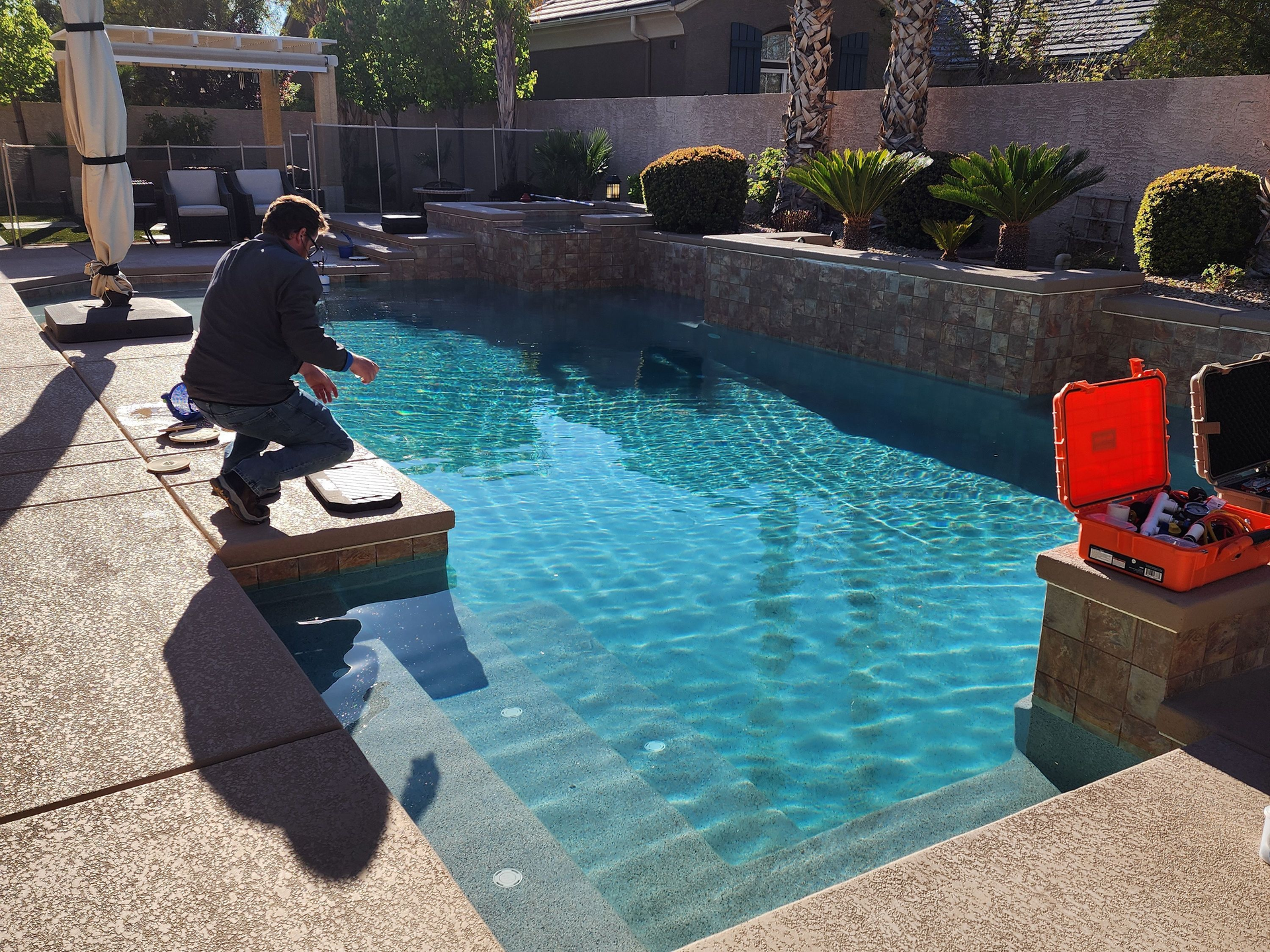 Person kneeling on a board beside a turquoise pool, with tools on the pool deck in bright sunlight
