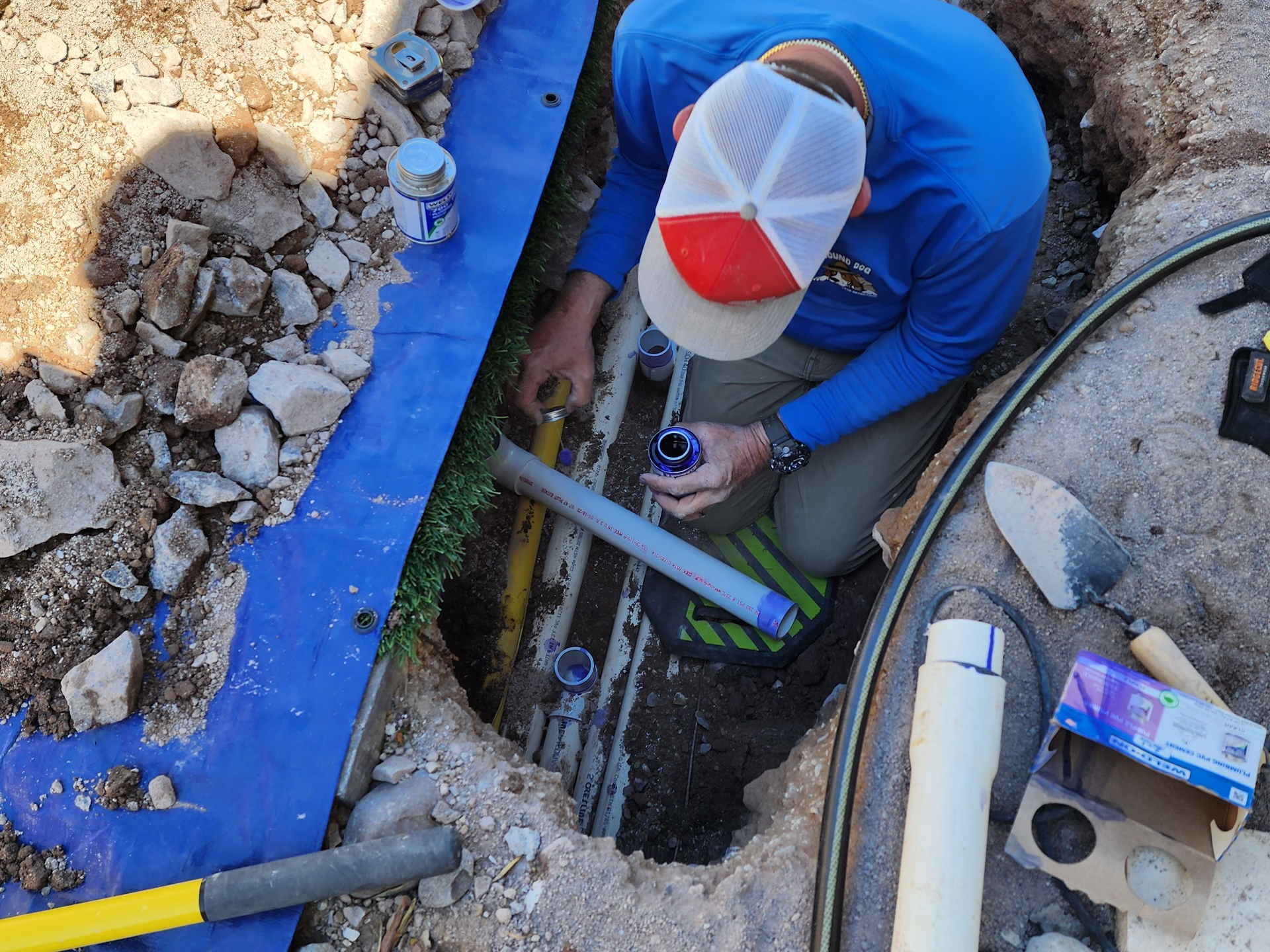 Worker in blue shirt repairing underground pipes in a trench with rubble and tools.