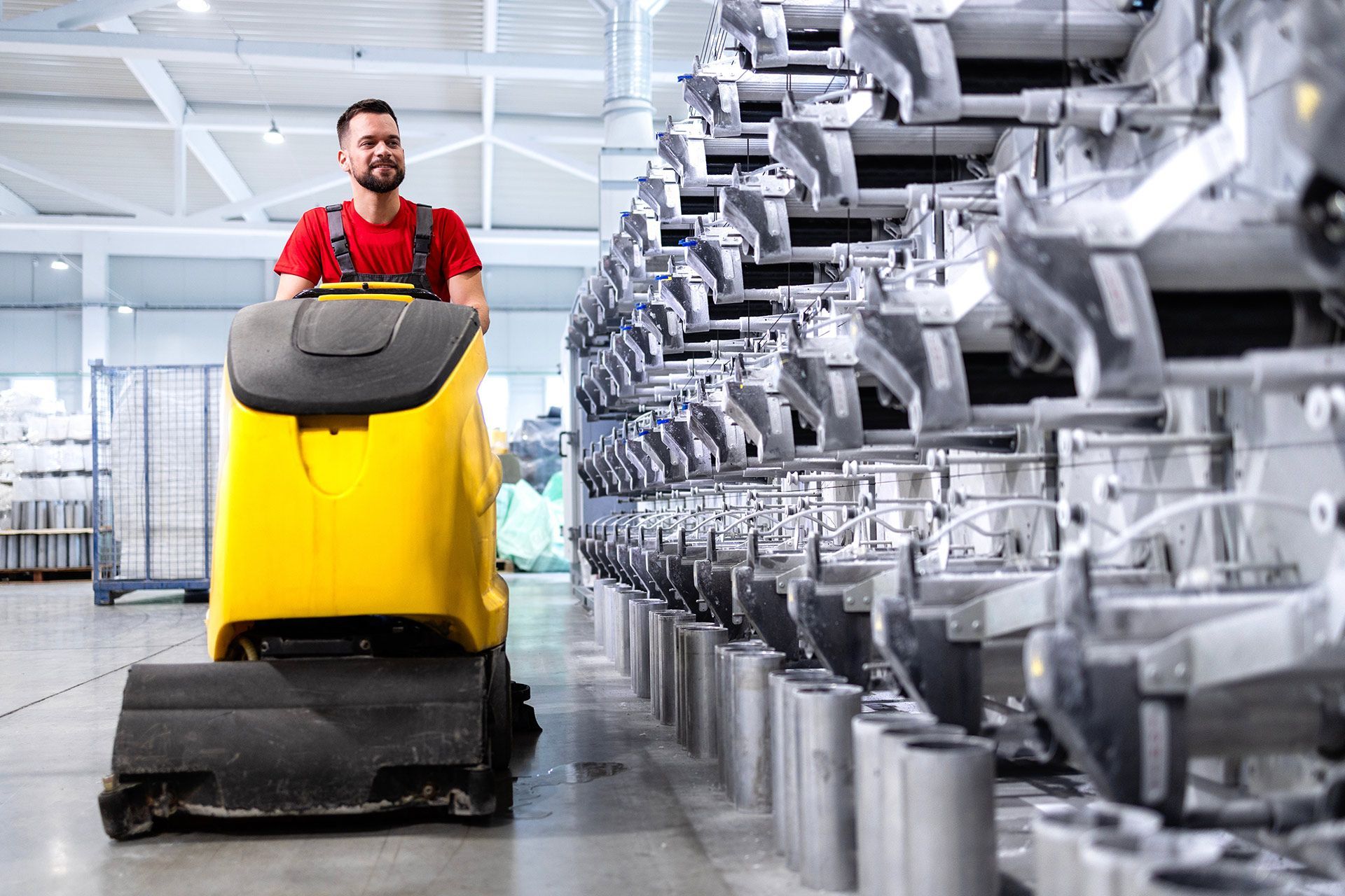 A person in a red shirt operates a yellow floor-scrubbing machine in a large warehouse aisle with metal racks.