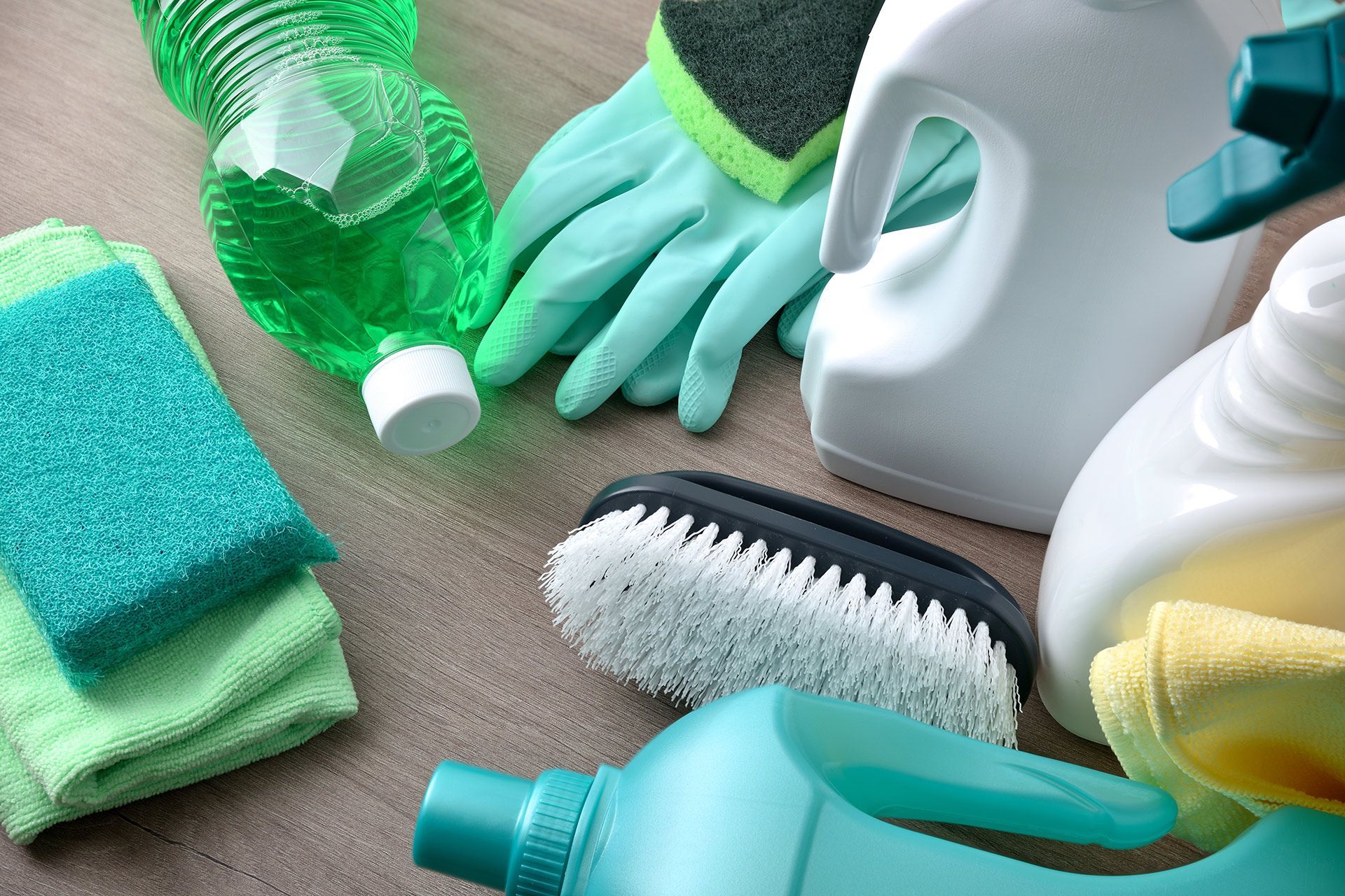 Cleaning supplies including bottles, brushes, gloves, and cloths arranged on a wooden surface.