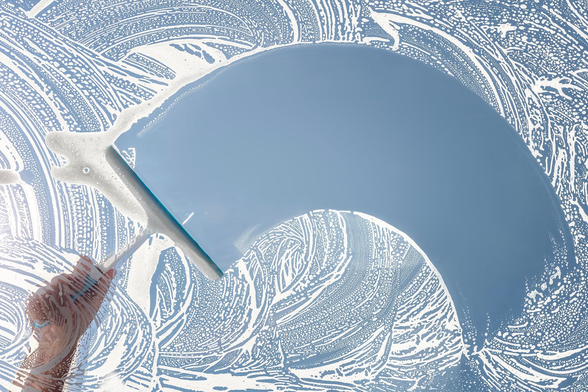 A squeegee held by a hand clears a streak of sudsy foam off a glass surface, revealing a clear blue sky underneath.