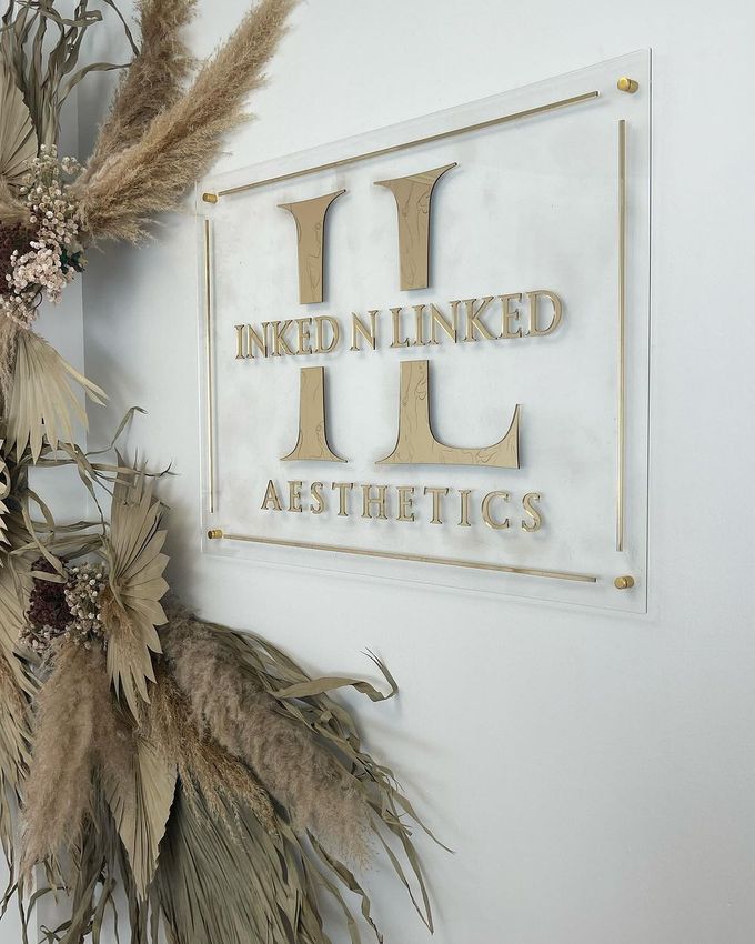 Gold “INKED N LINKED AESTHETICS” sign on a white wall with dried pampas grass decor