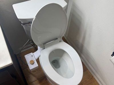 White toilet with the seat up, a bidet attachment, and a control panel are in a bathroom