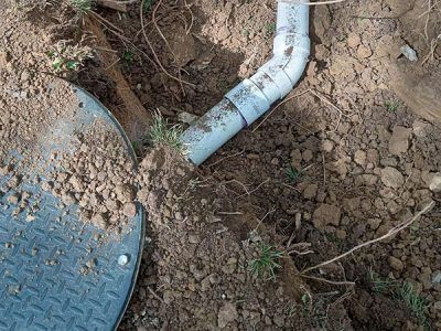 A section of buried PVC pipe emerging from the dirt next to a metal access cover.