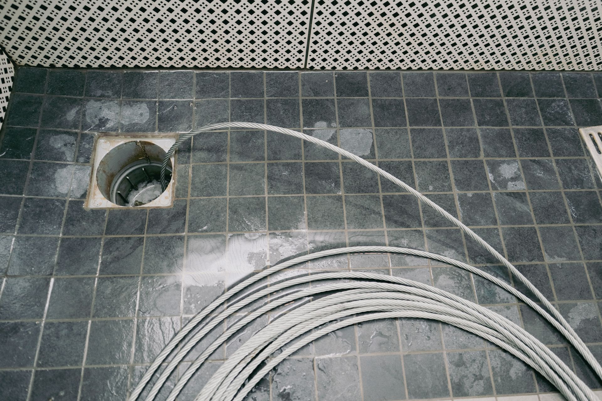 Gray coiled tubing extended into a drain on a tiled floor with overhead grating.