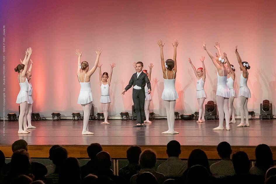 Des danseurs en blanc et un autre en noir évoluent sur une scène dont le fond est éclairé en rose.