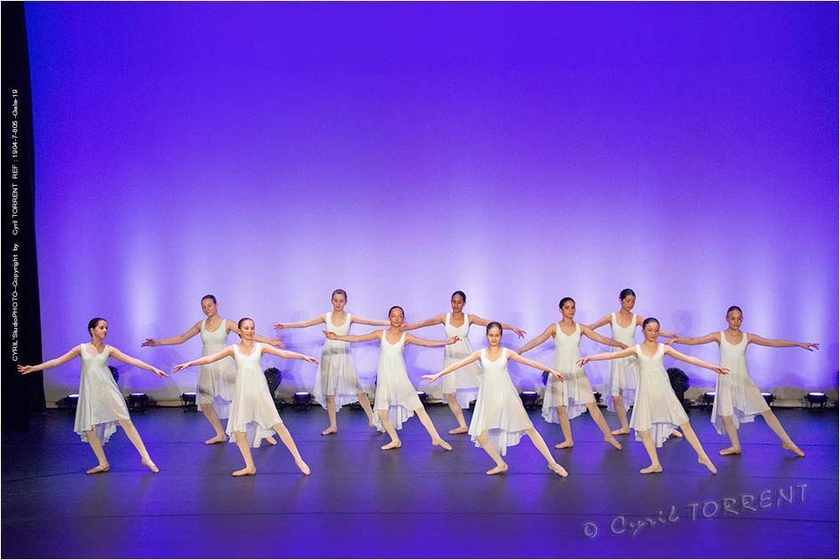 Des danseurs de ballet en costumes blancs exécutent une danse en ligne synchronisée sur une scène éclairée en bleu