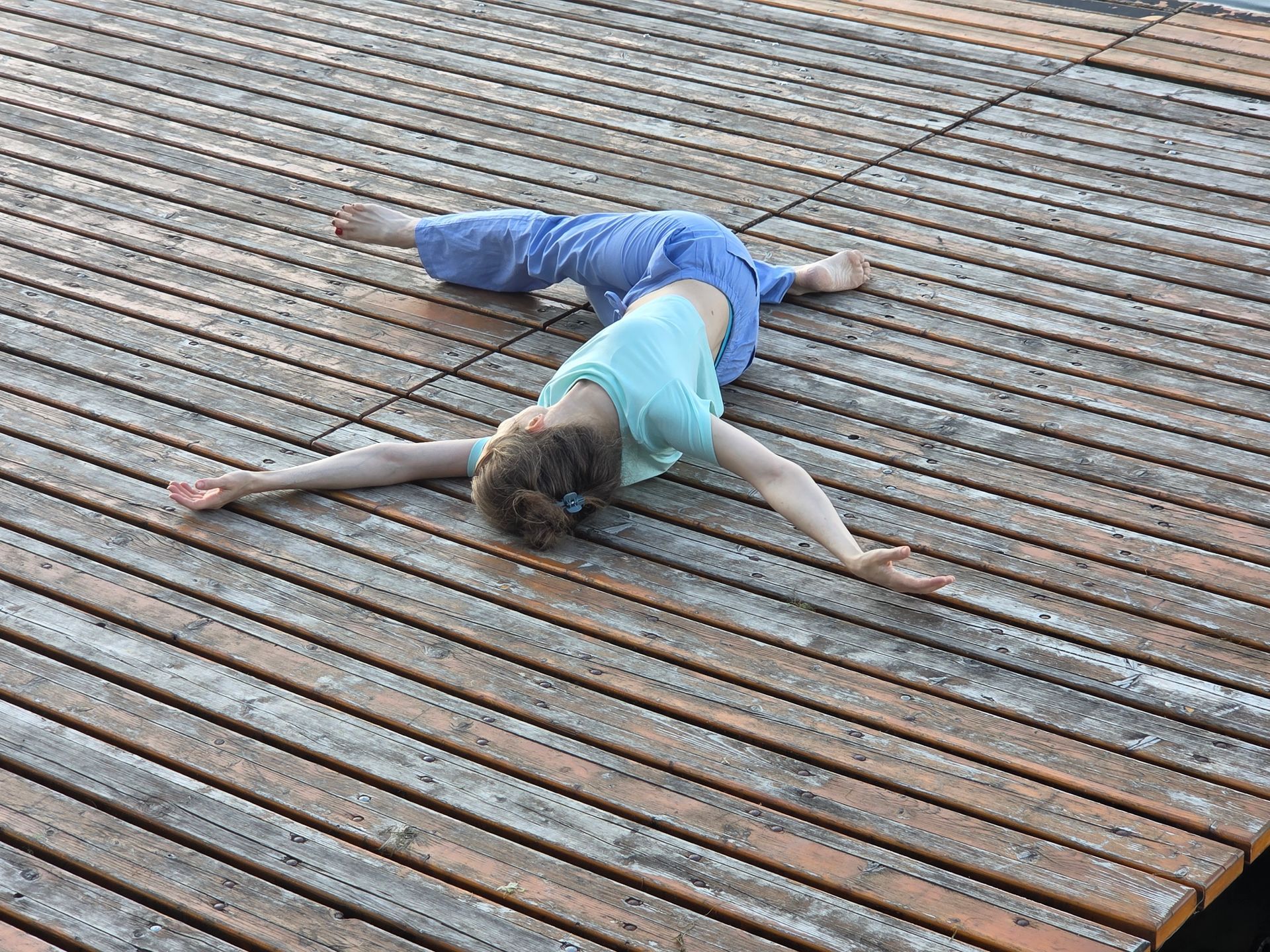 Personne allongée à plat ventre sur une terrasse en bois, portant un haut bleu clair et un pantalon bleu