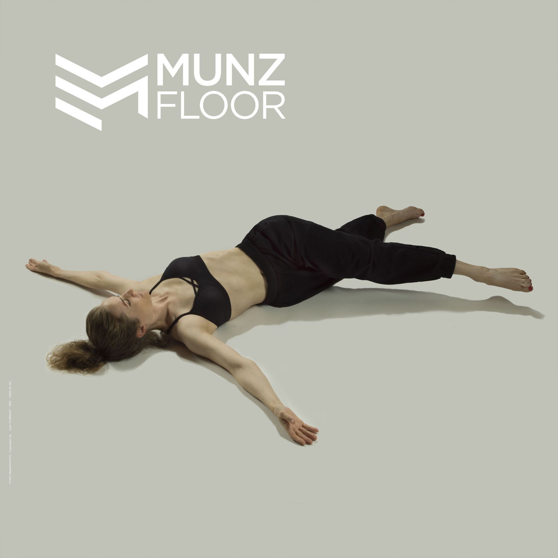 Femme en tenue de sport noire allongée sur un sol gris clair, avec le logo Munz Floor dans un coin.