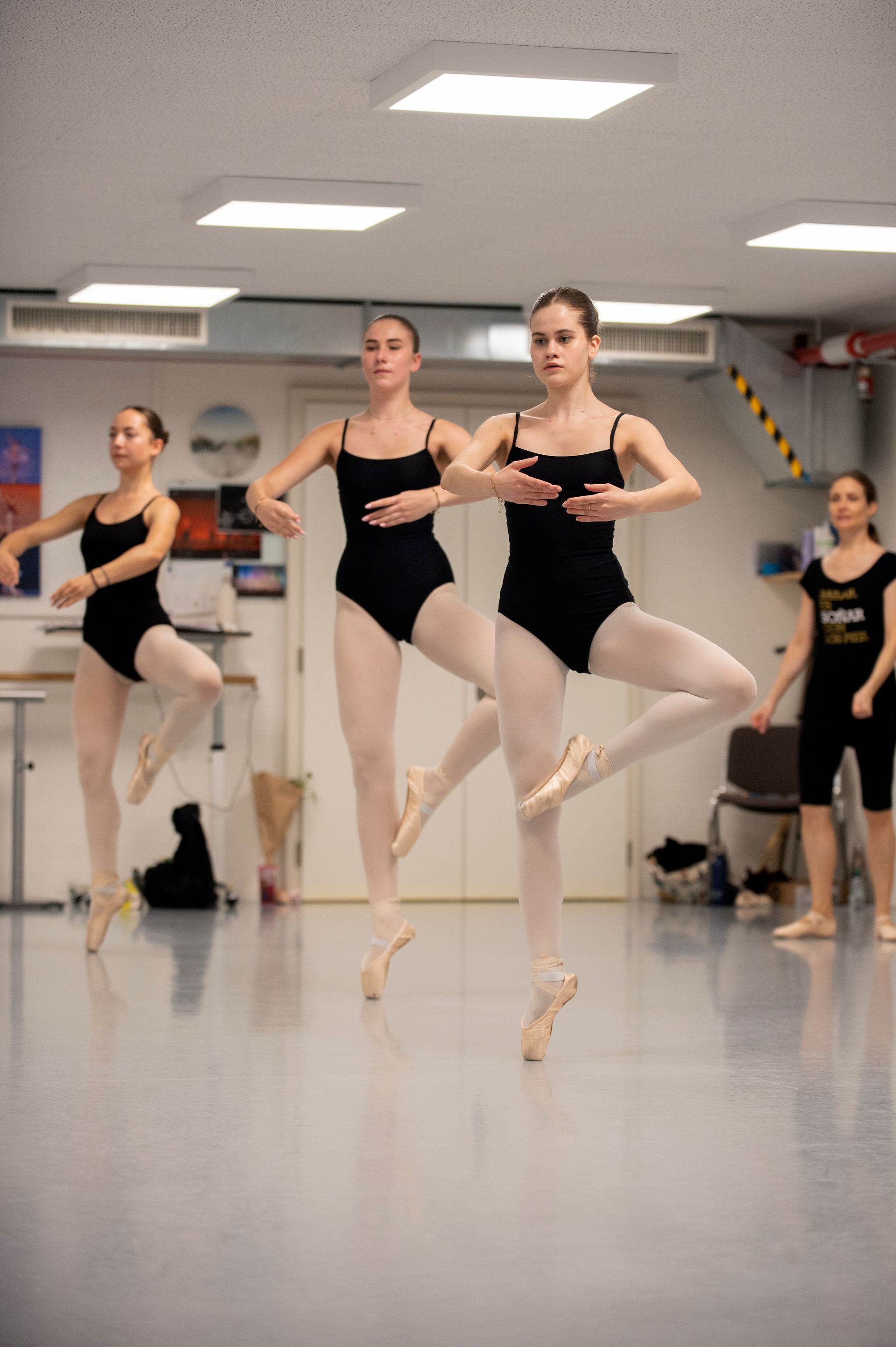 Des danseuses de ballet en justaucorps noirs s'entraînent en studio, en équilibre sur pointes, les bras levés.