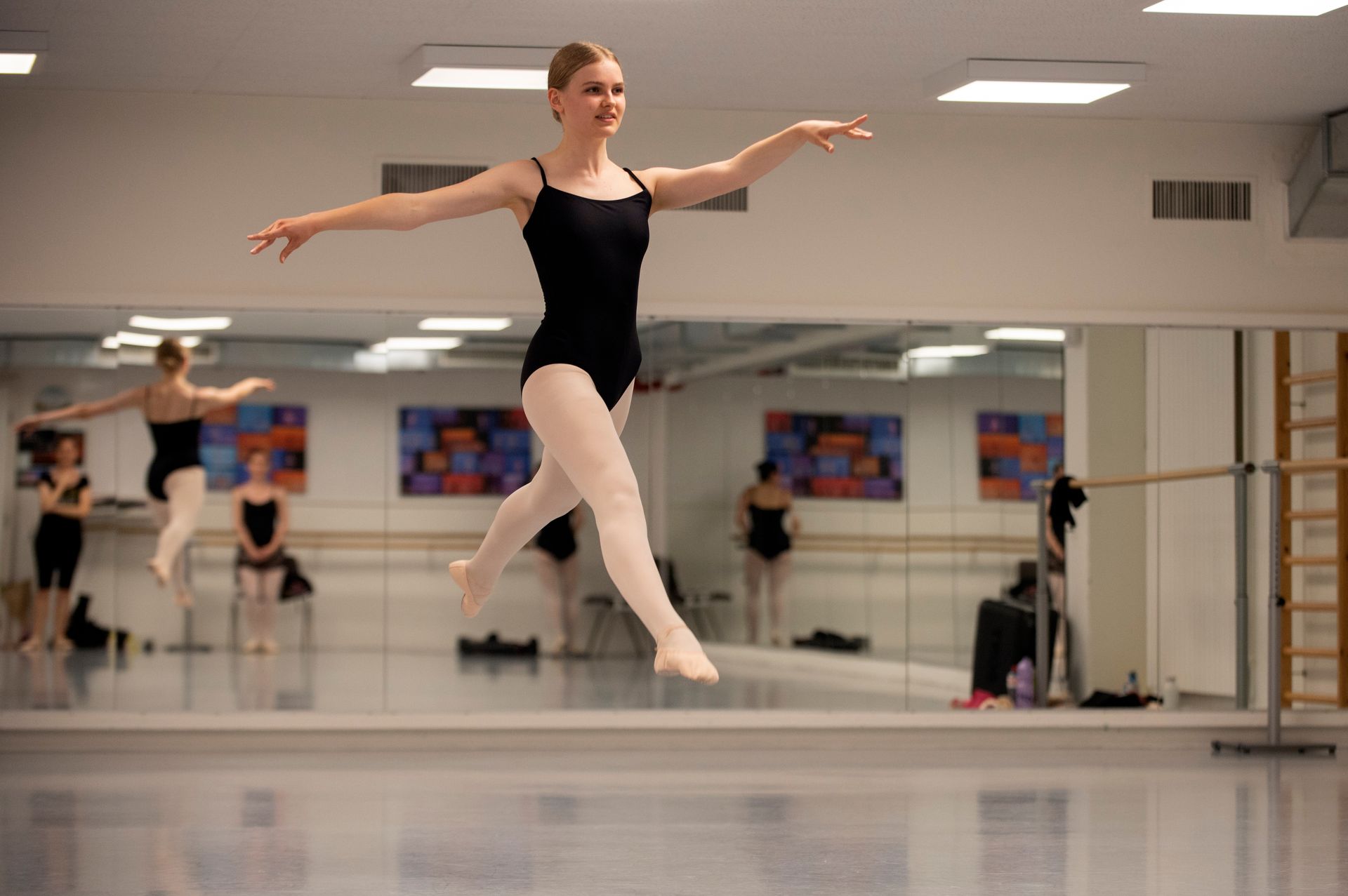 Une danseuse de ballet en justaucorps noir et collants roses s'entraîne à un saut dans un studio avec des miroirs.