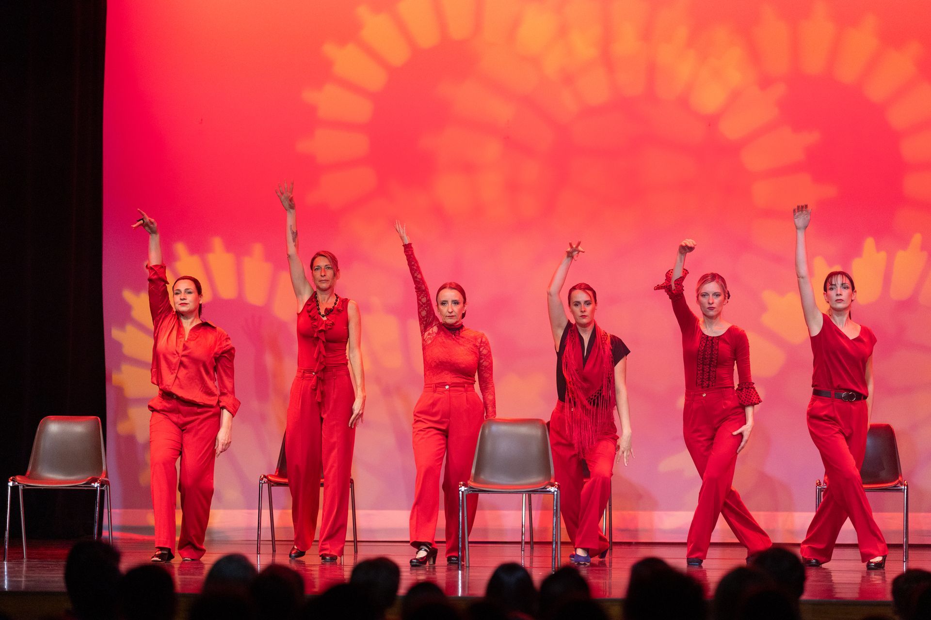Six artistes vêtus de rouge posent les bras levés sur une scène avec des chaises et un fond rouge.