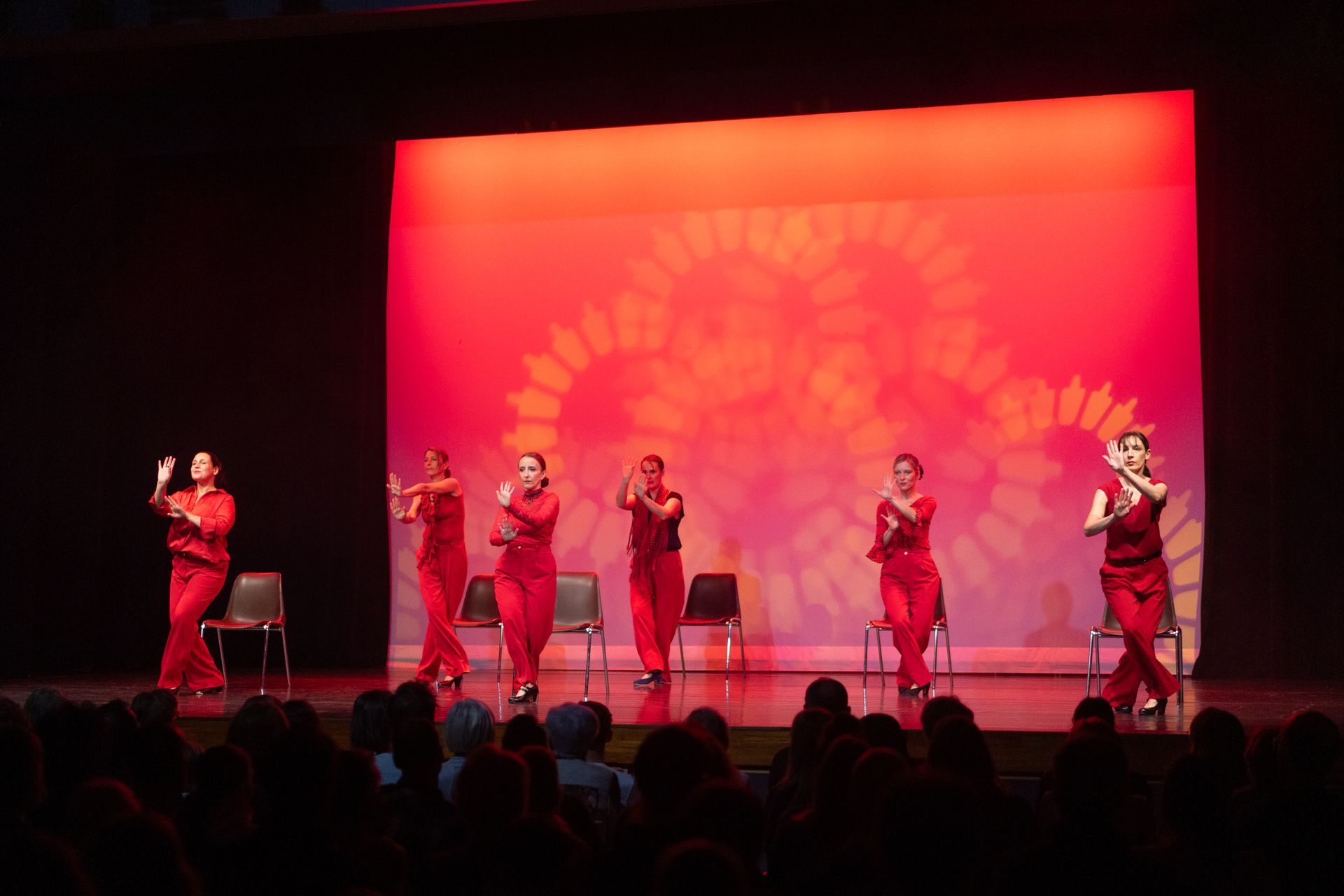 Six artistes vêtus de rouge dansent sur une scène éclairée de rouge devant un public