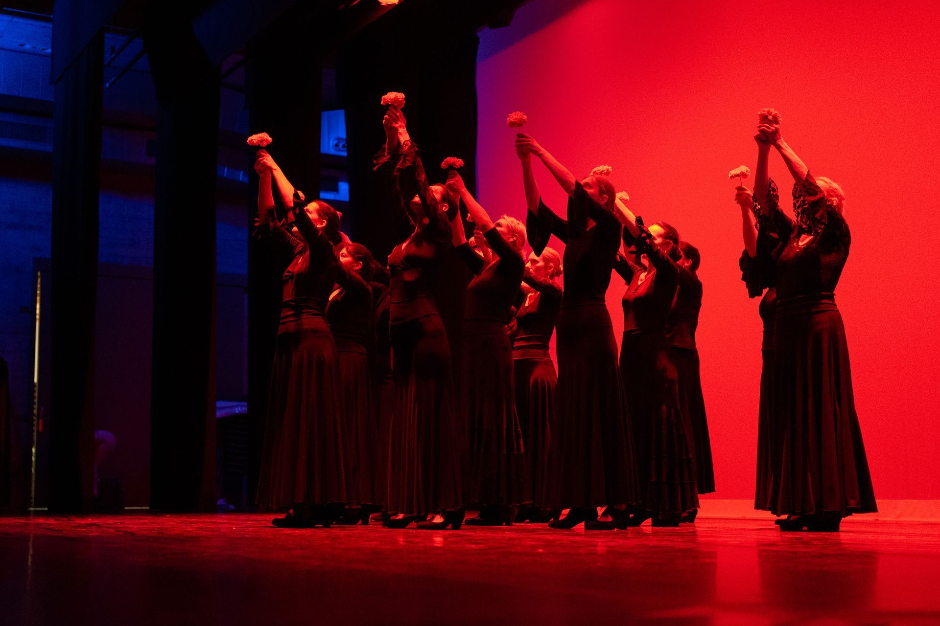 Des danseuses en longues robes noires, les bras levés, évoluent sous un éclairage de scène rouge dramatique.