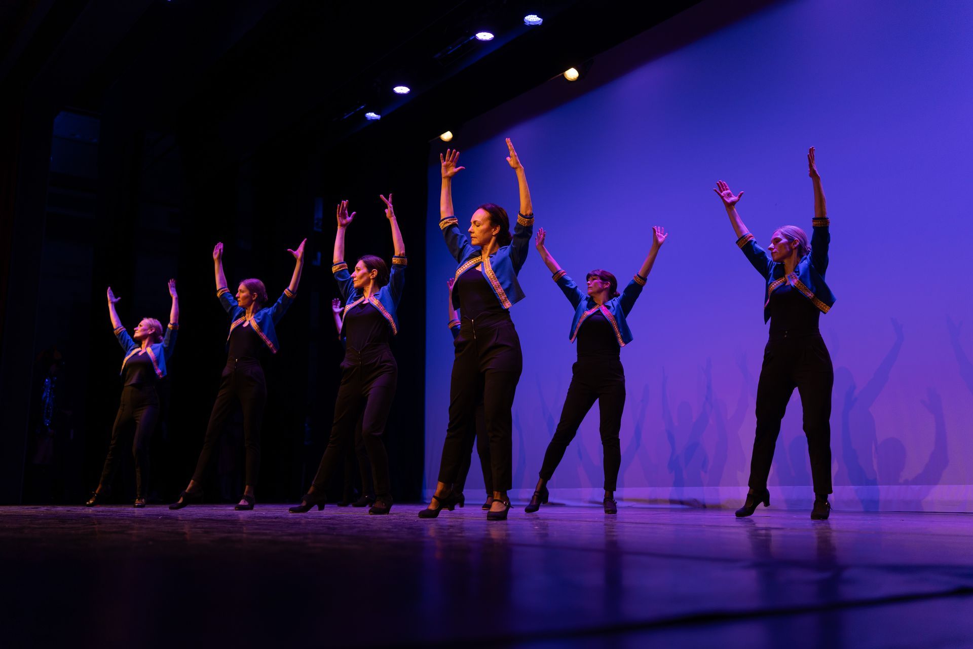 Des danseurs en costumes noirs, les bras levés, sous une lumière de scène violette