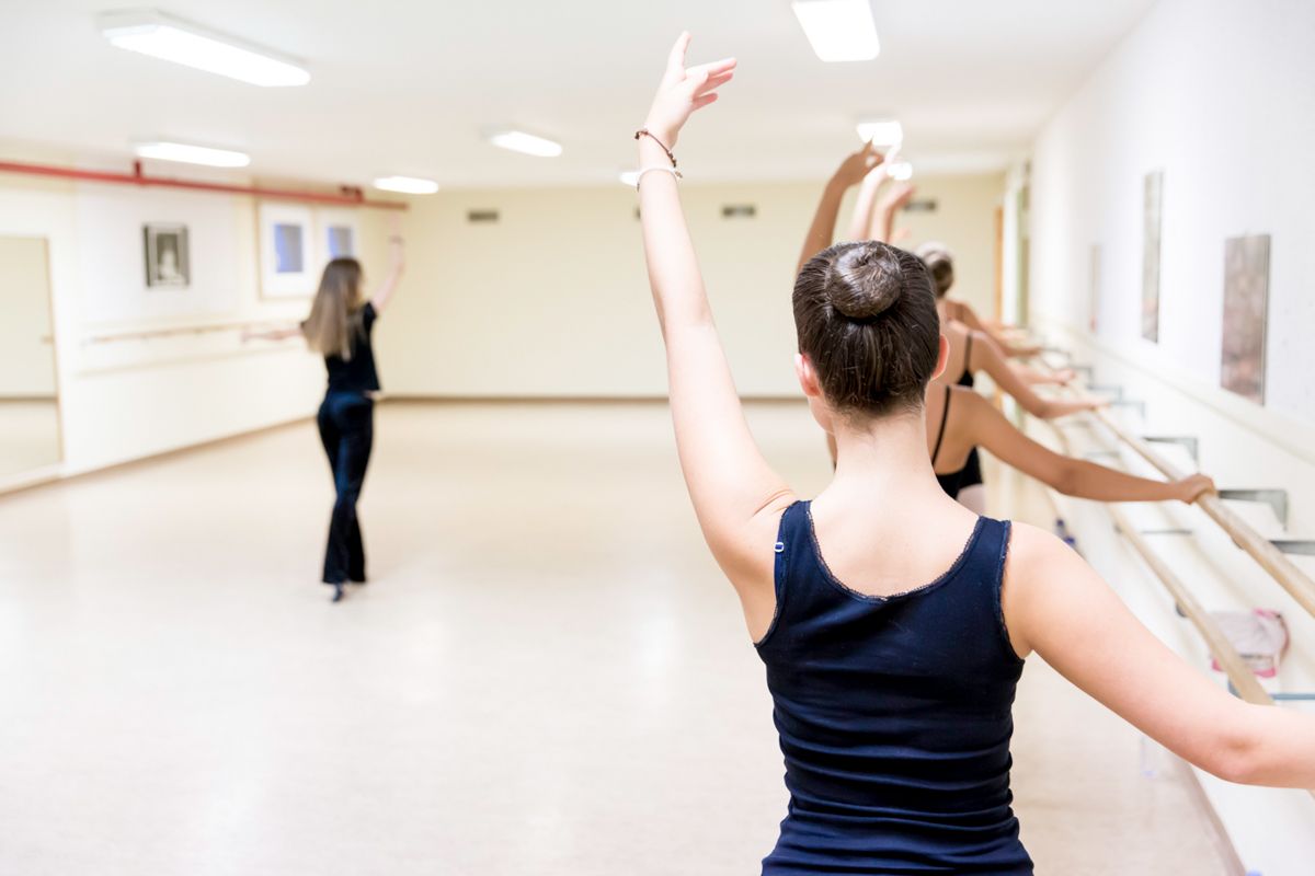 Des danseuses de ballet s'entraînent dans un studio lumineux, l'une d'elles au premier plan, les bras levés, des miroirs le long du mur.