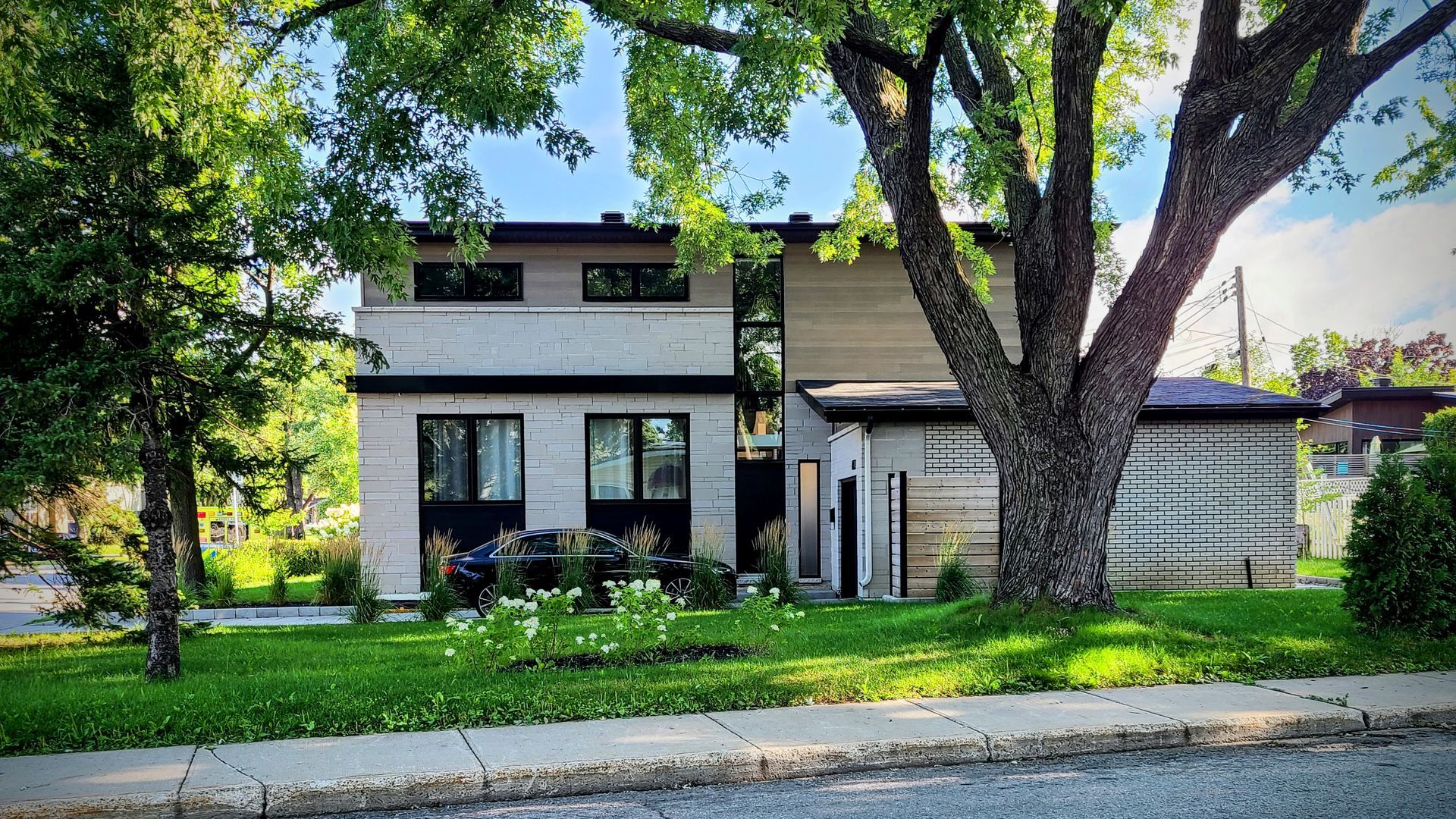 Maison à deux étages avec façade en briques claires et bardage en bois, arbres devant, pelouse verte.