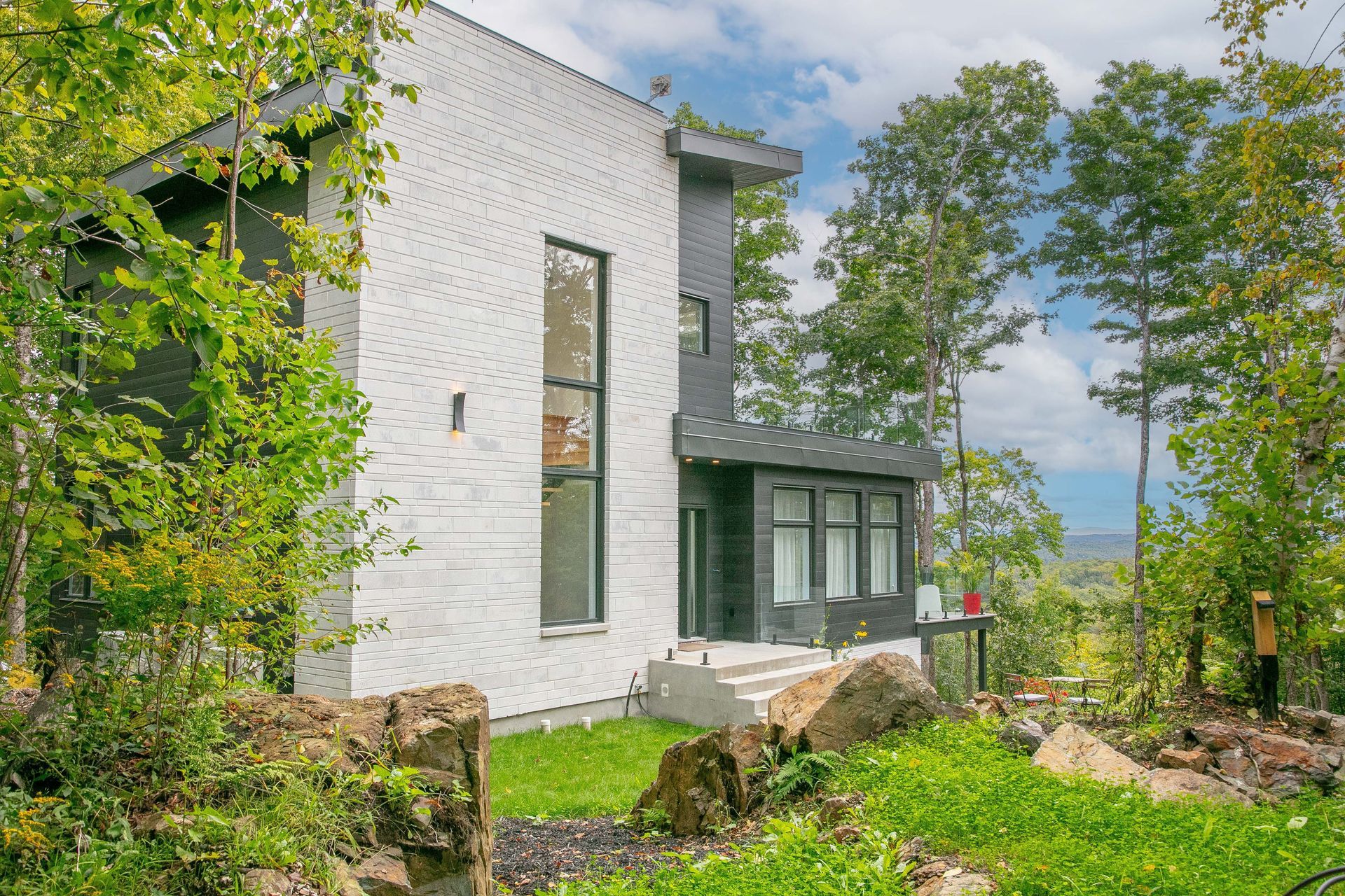 Maison moderne blanche et grise avec de grandes fenêtres, nichée au milieu des arbres à flanc de colline.