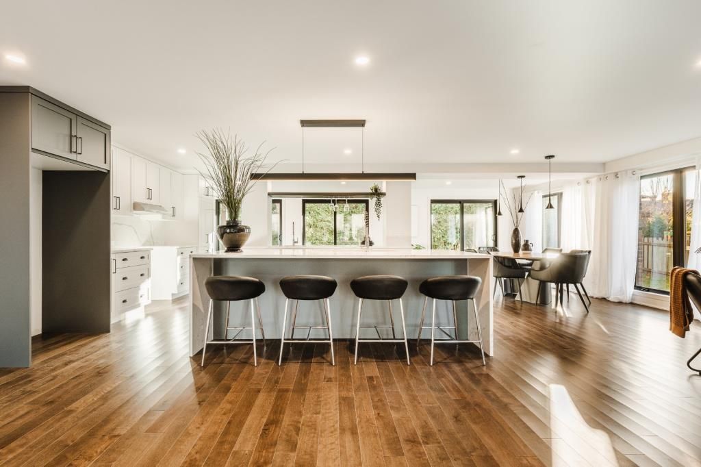 Cuisine moderne avec îlot central, tabourets de bar et coin repas avec grandes fenêtres et parquet.