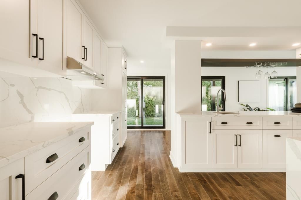 Cuisine blanche avec quincaillerie foncée, crédence en marbre et parquet. Porte-fenêtre coulissante donnant sur l'extérieur.