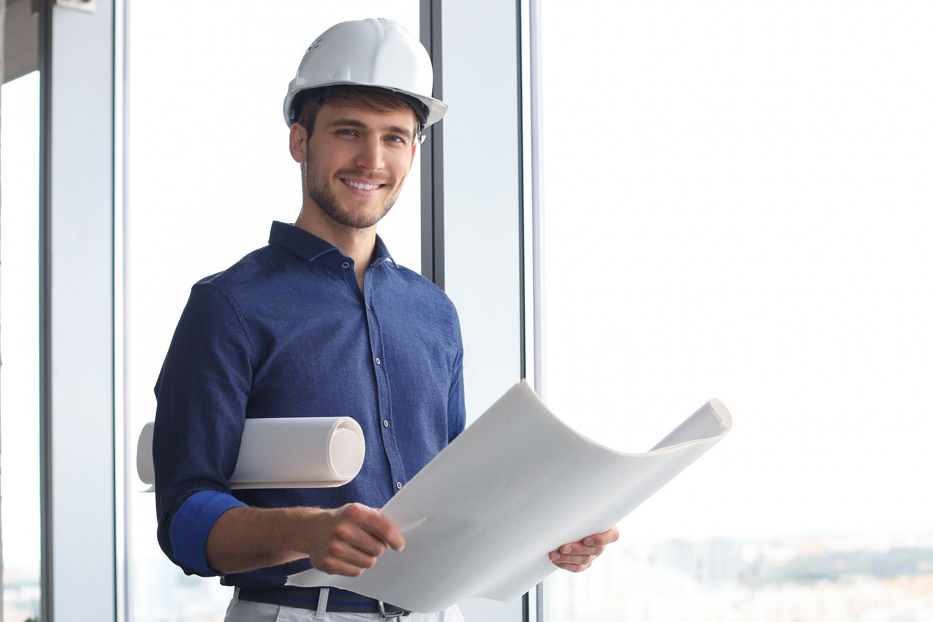 Un ouvrier du bâtiment, casque de chantier sur la tête, sourit en tenant des plans près d'une fenêtre.