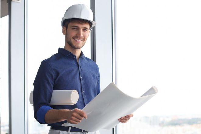 Un ouvrier du bâtiment, casque de chantier sur la tête, sourit en tenant des plans près d'une fenêtre.