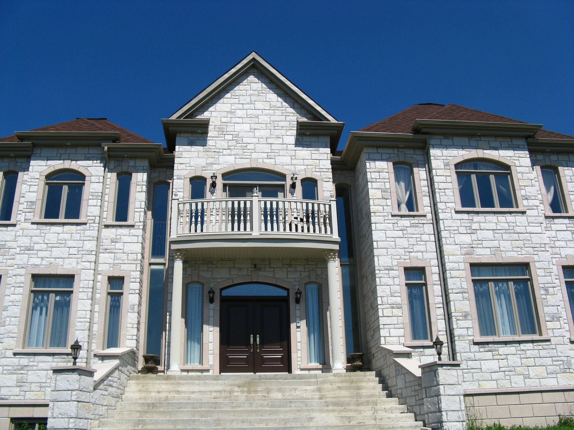 Maison à deux étages avec façade en pierre, portes sombres et balcon, sous un ciel bleu limpide.