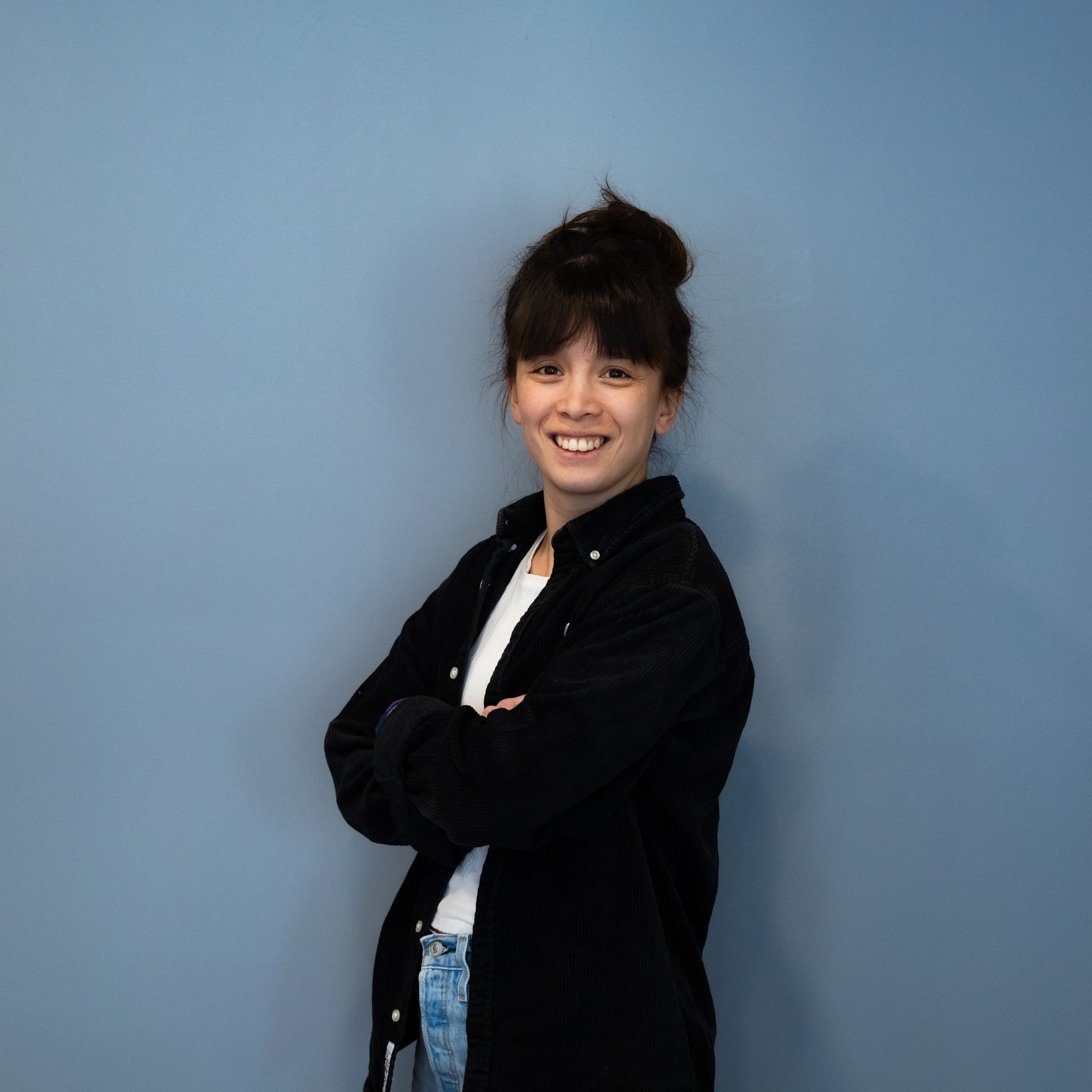 Femme les bras croisés, souriante, vêtue d'un t-shirt noir et d'un jean, devant un mur bleu.