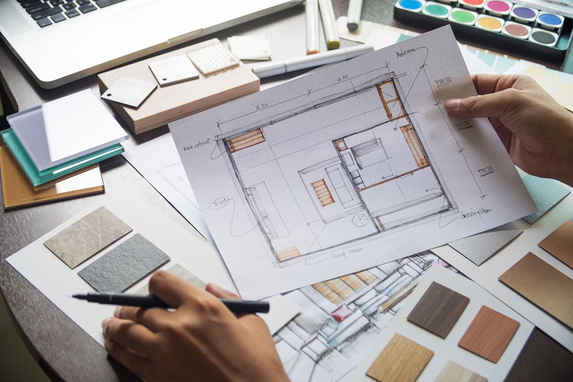 Des mains tenant un plan d'architecte, entourées d'outils de conception et d'échantillons de couleurs.