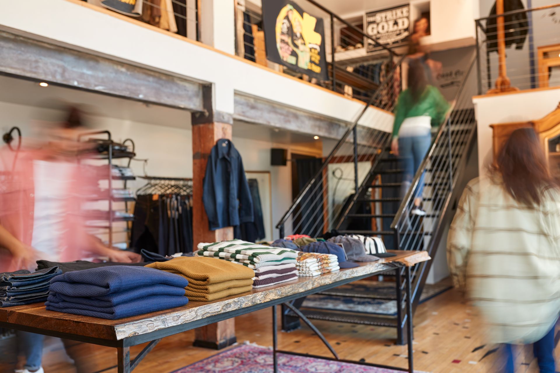 Intérieur d'un magasin de vêtements avec des clients qui font leurs achats. Table d'exposition avec des pulls pliés, escalier menant au deuxième étage.