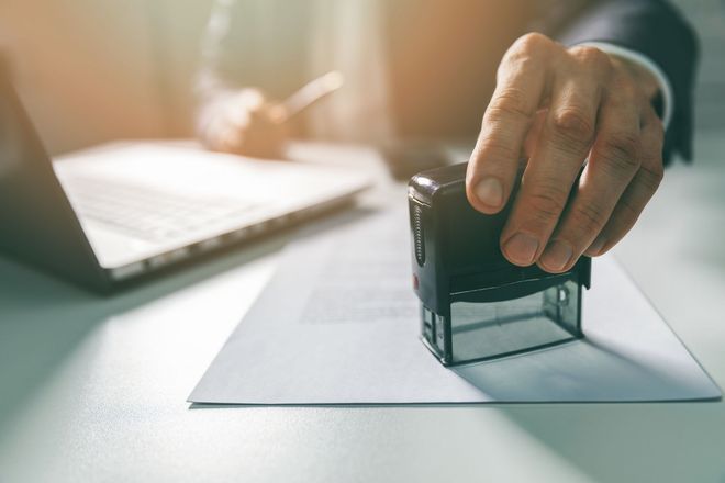 Une personne en costume appose un tampon encreur automatique sur un document, assise à un bureau ; un ordinateur portable se trouve à proximité.