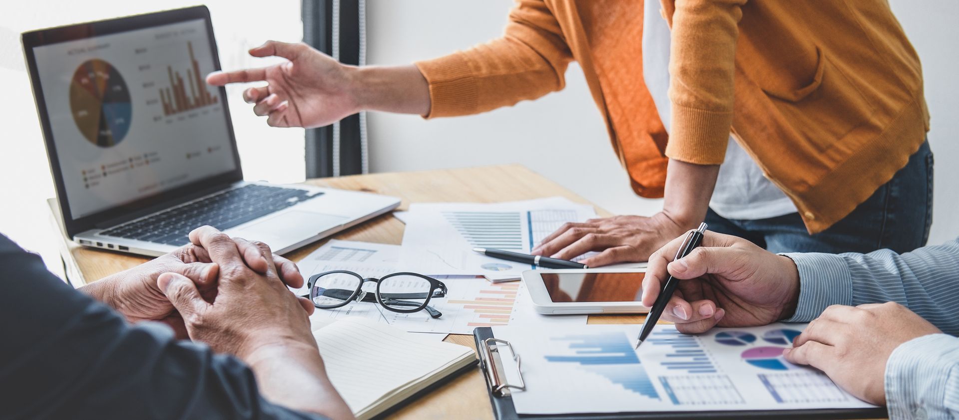 Des personnes analysent des données, l'une d'elles désignant l'écran d'un ordinateur portable affichant un graphique circulaire. Des papiers, une tablette et des lunettes sont posés sur le bureau.
