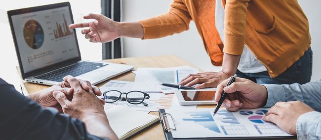 Des personnes analysent des données, l'une d'elles désignant l'écran d'un ordinateur portable affichant un graphique circulaire. Des papiers, une tablette et des lunettes sont posés sur le bureau.