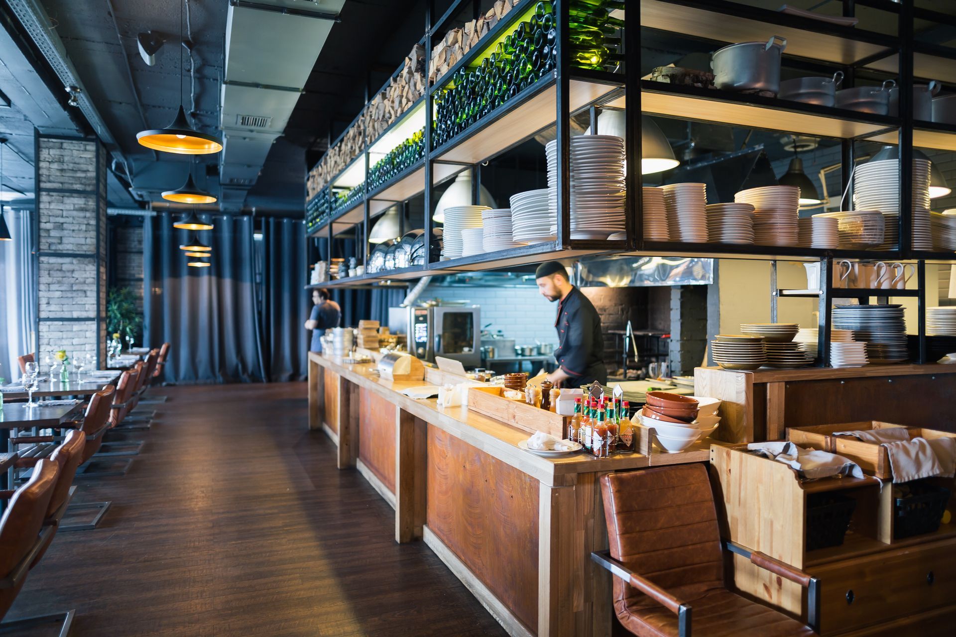 Intérieur d'un restaurant avec un chef au travail, une cuisine ouverte, des tables à manger et des étagères suspendues.