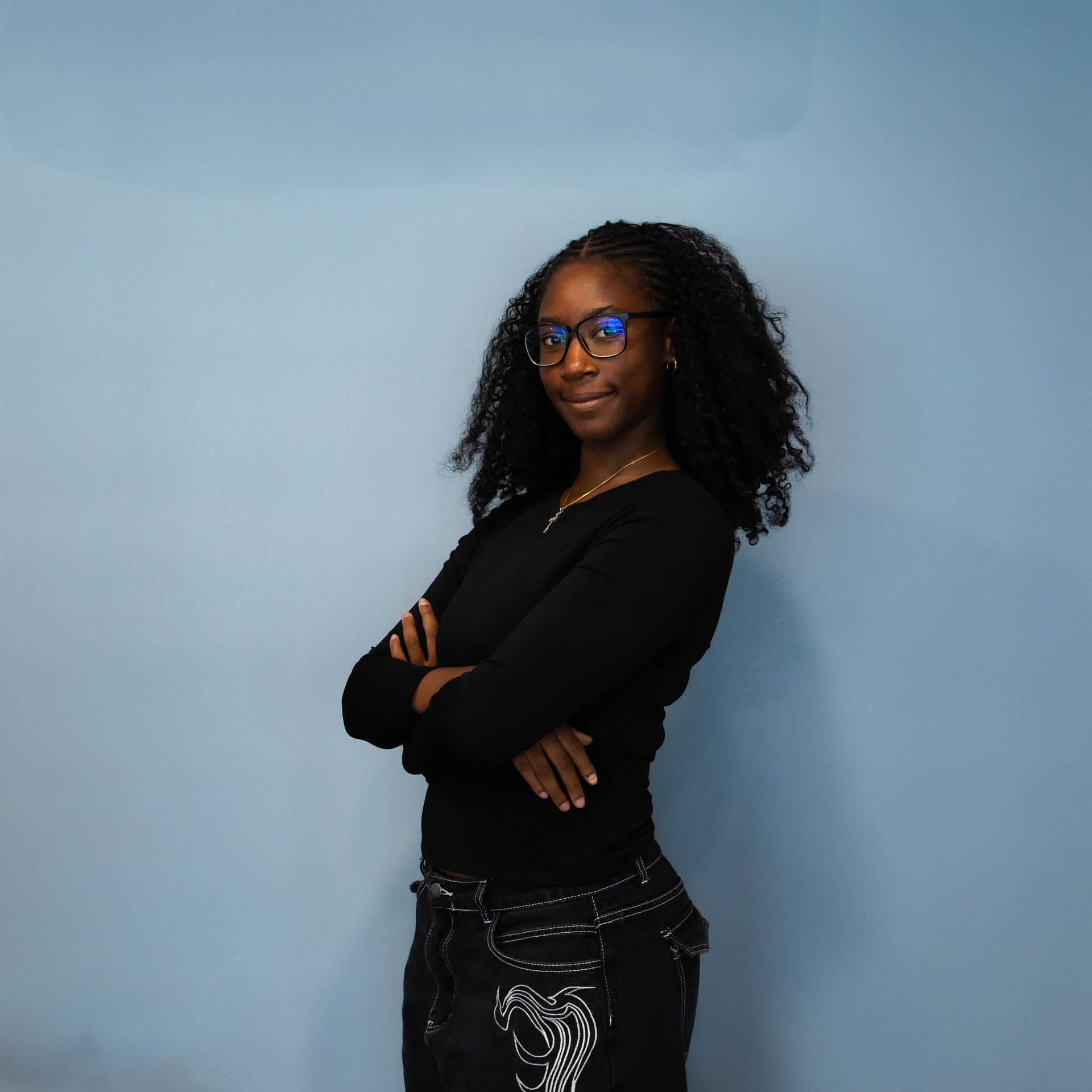 Femme à lunettes, bras croisés, souriante. Vêtue d'un haut noir et d'un jean, elle se détache sur un fond bleu.