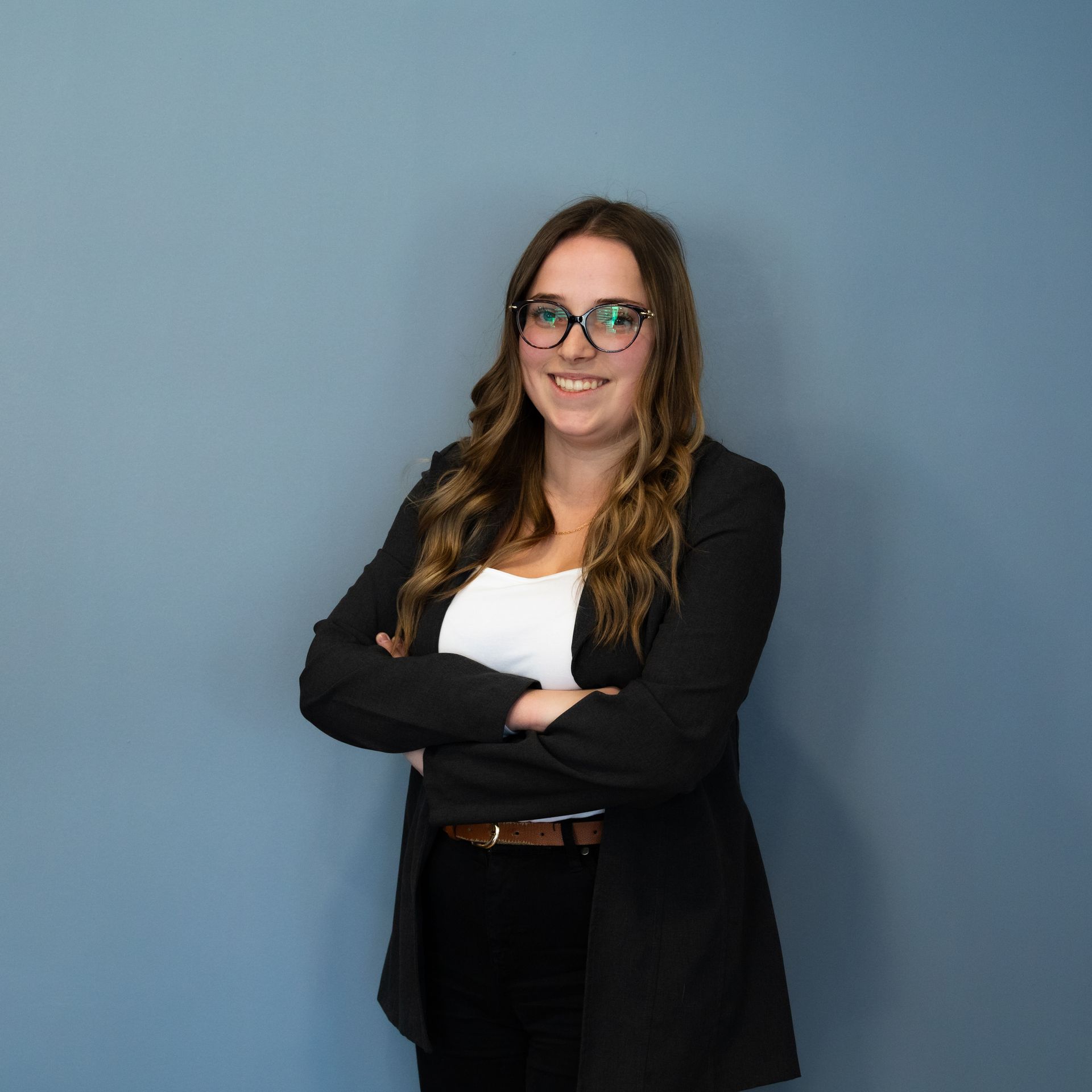 Femme portant des lunettes et un blazer, les bras croisés, souriante. Fond de mur bleu.