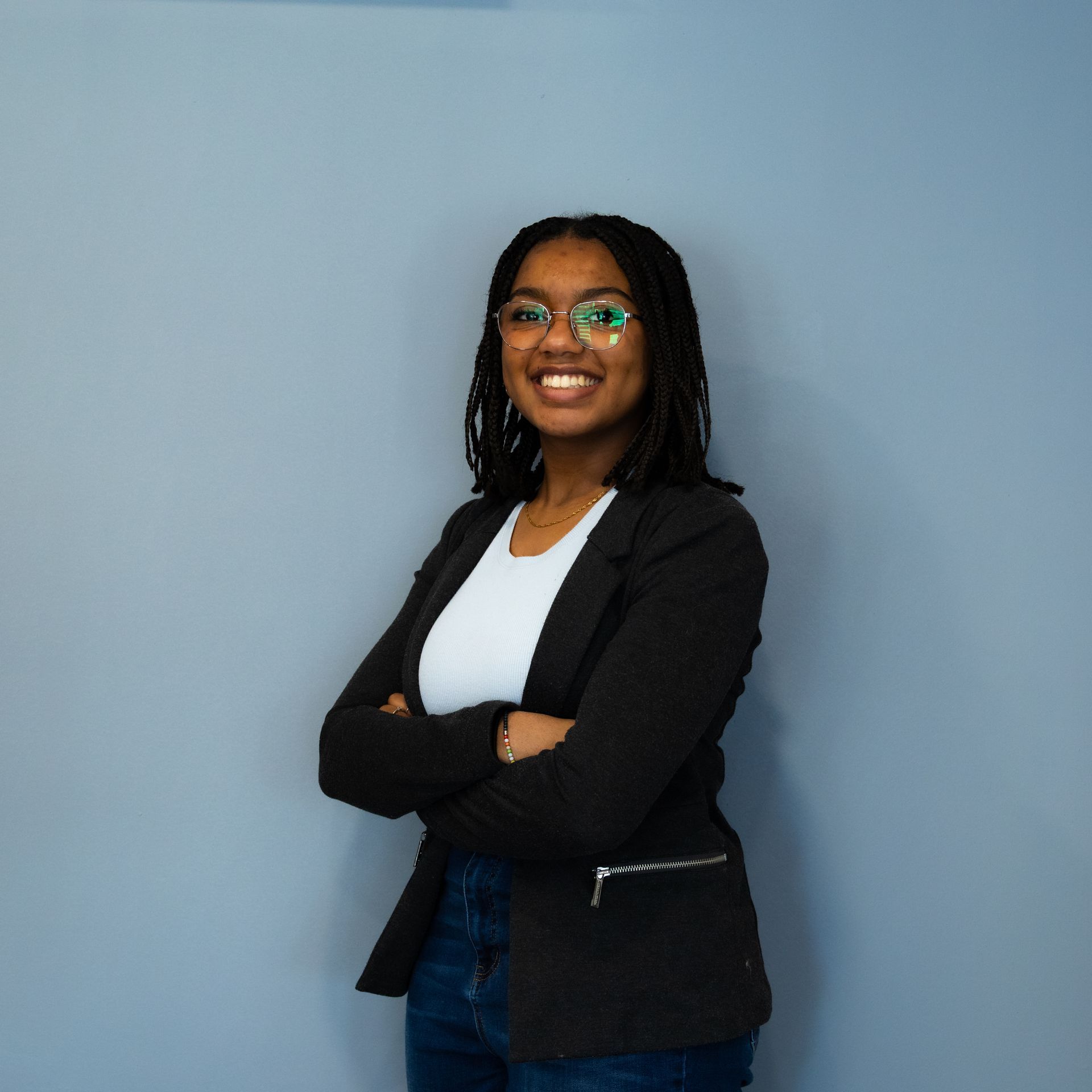 Femme à lunettes, bras croisés, souriante. Elle porte un blazer, une chemise blanche et un jean devant un fond bleu.