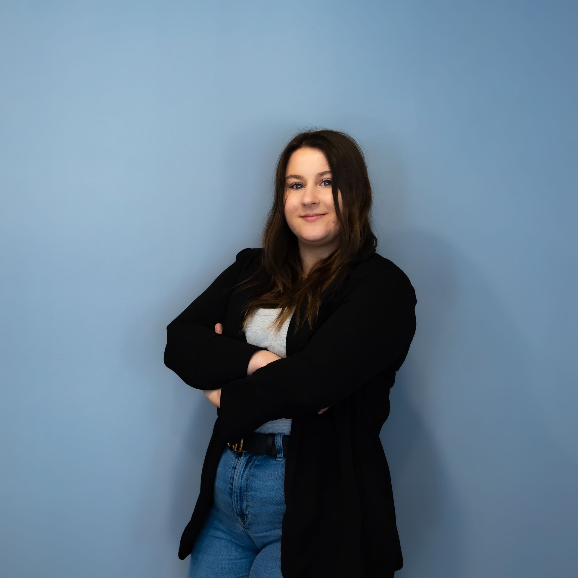 Femme les bras croisés, vêtue d'un jean, d'un blazer noir et d'une chemise grise, devant un mur bleu.