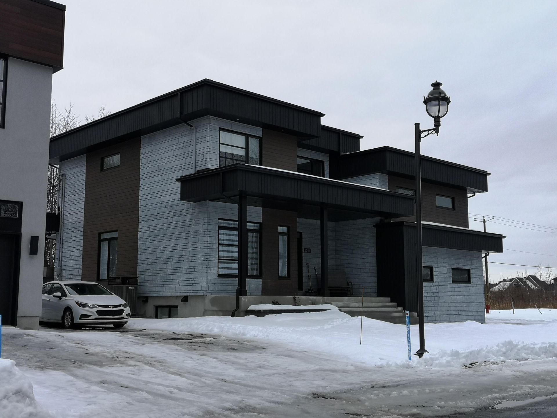 Maison moderne à deux étages avec façade en briques grises et abri voiture, dans un décor enneigé. Une voiture est garée devant.