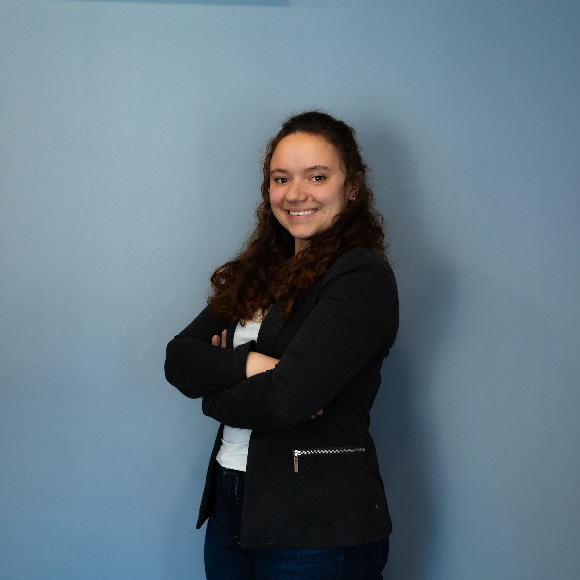 Une femme, les bras croisés, sourit devant un mur bleu ; elle porte un blazer noir et une chemise blanche.