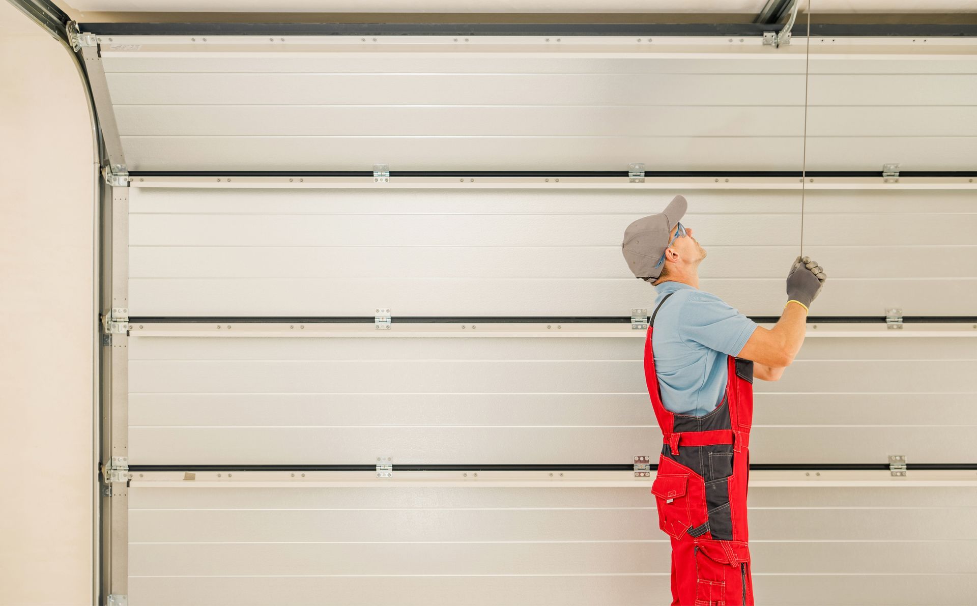 Man in red overalls repairing a white garage door, looking up, indoors.