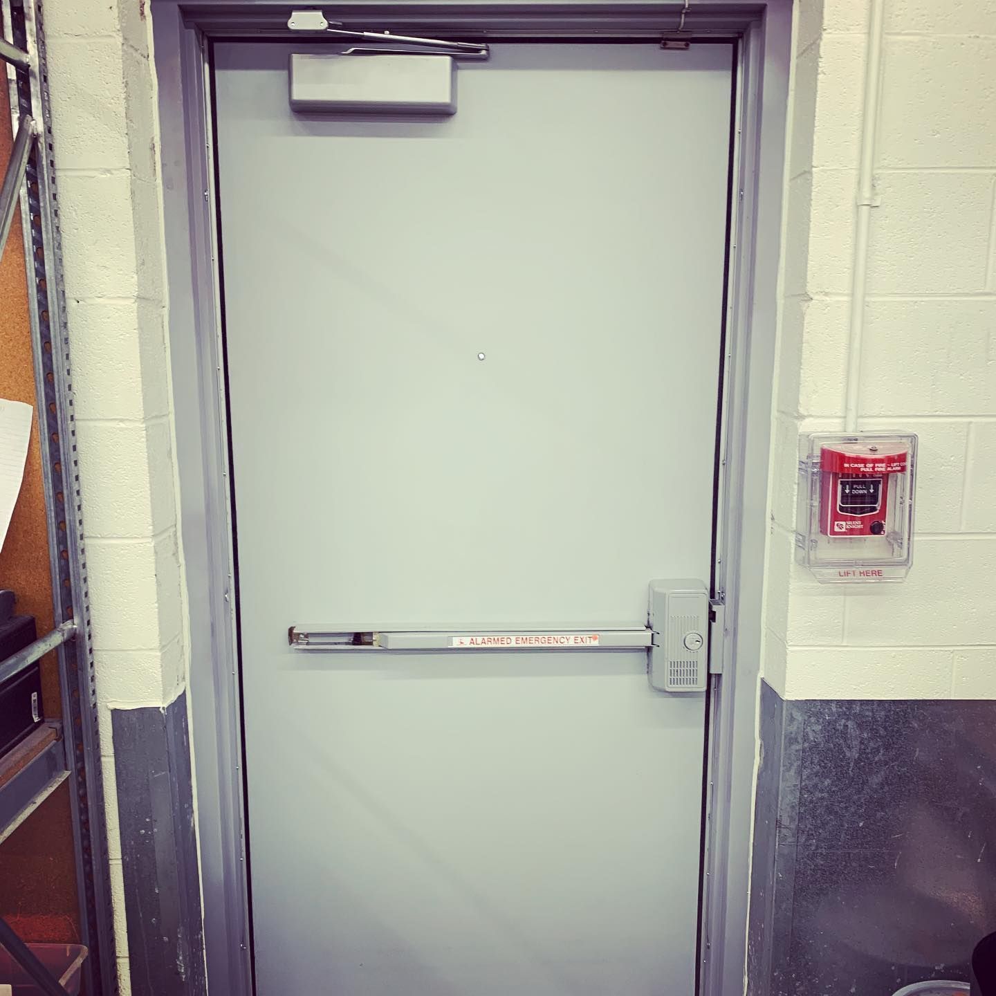 Metal fire door with push bar, near a fire alarm on a light-colored wall.