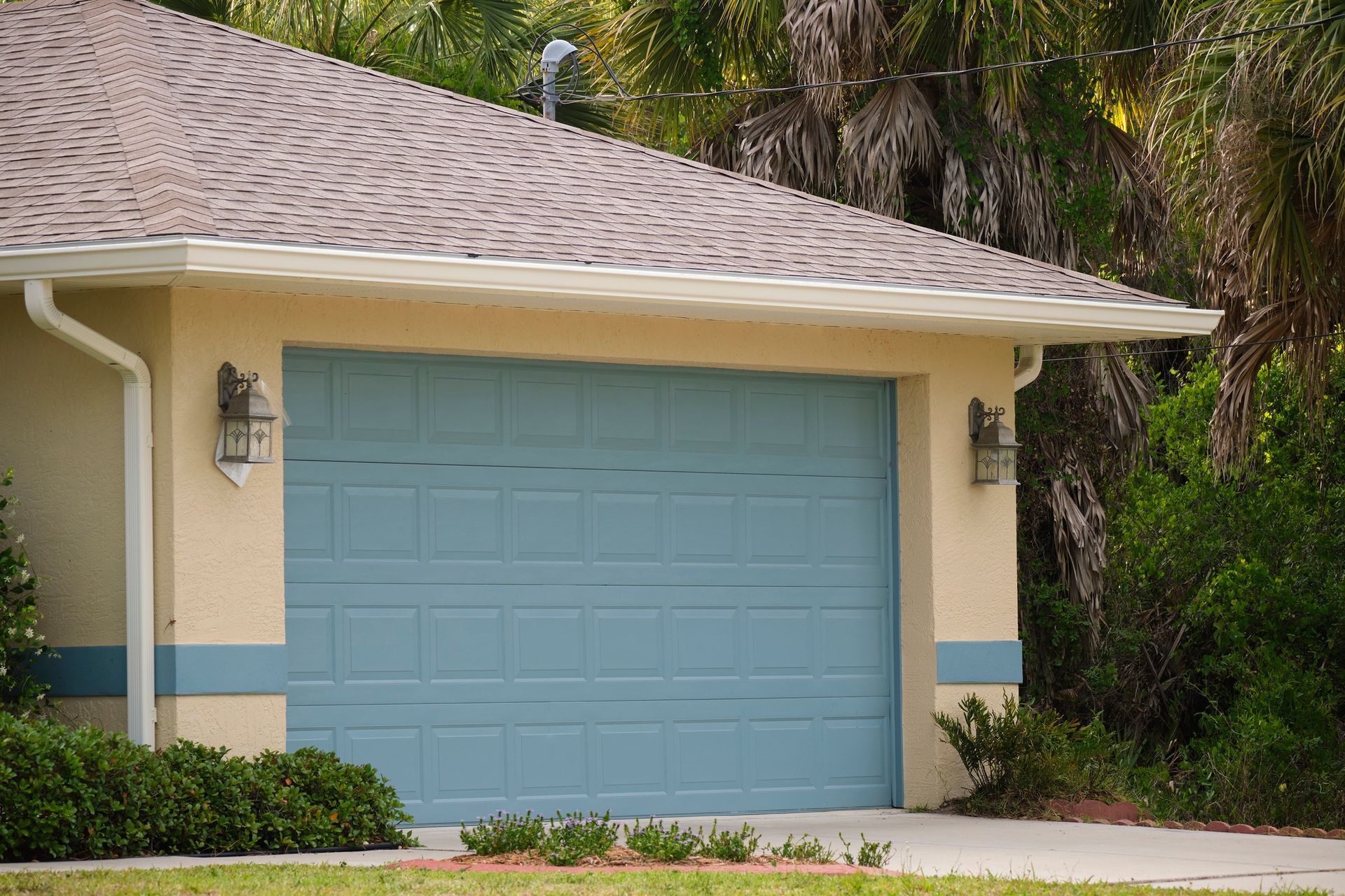 Blue garage door on a light yellow house with a brown roof and bushes.