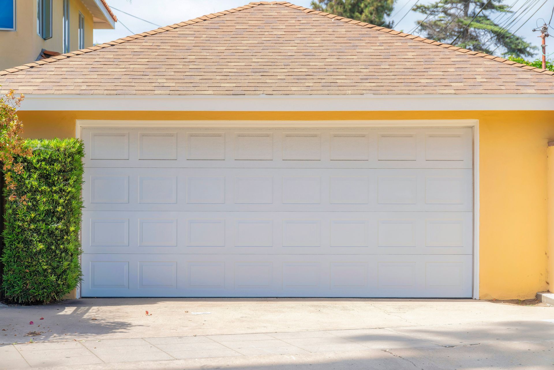 Newly Installed Garage Door — Detroit, MI — Town & Country Door White garage door on a yellow building, with brown roof and green bush.
