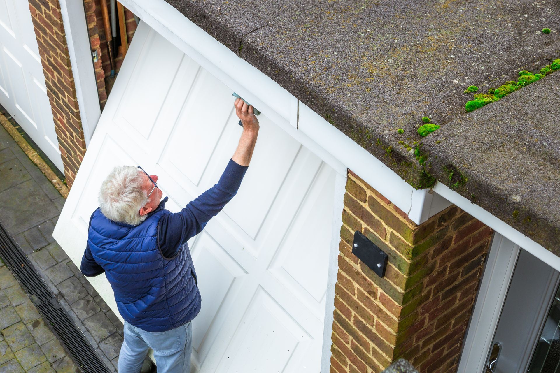 Man opening a white garage door, seen from above. Brick and concrete building, moss on roof.