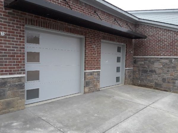 Two light gray garage doors with square glass panels, brick building exterior. Concrete driveway, dark awning.