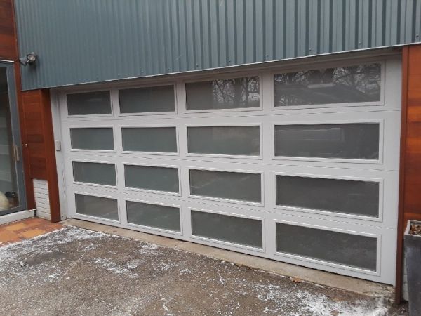 A light gray garage door with rectangular glass panels, set in a building with wood and gray siding.