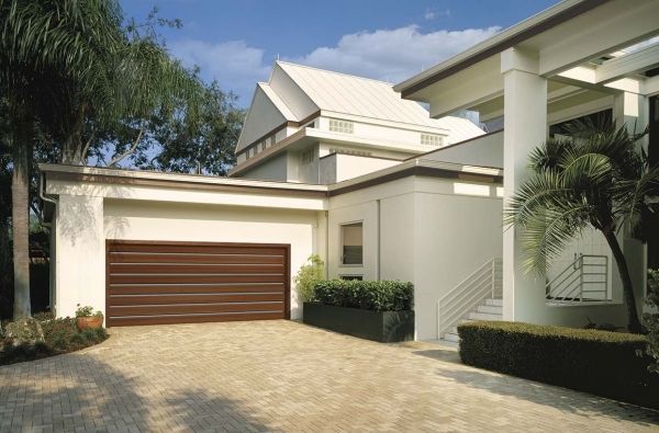 Brown garage door on a beige house with a brick driveway and palm trees.