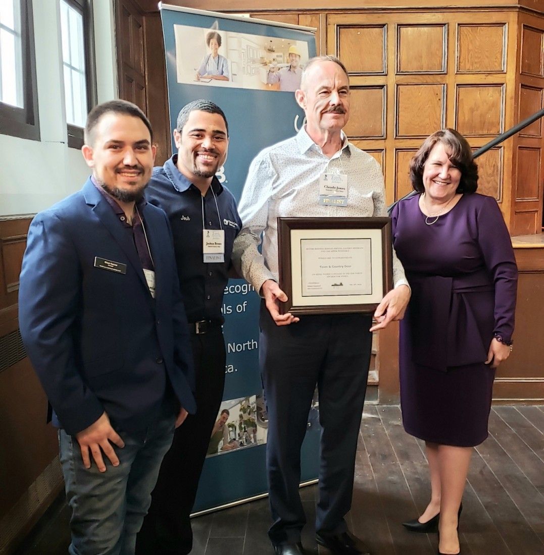 Four people pose with an award in a wood-paneled room. Man holds framed certificate, others smile. A banner is behind them.