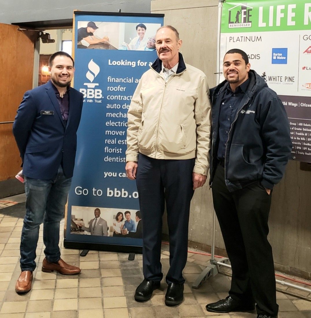 Three men stand near a Better Business Bureau banner. One wears a blue blazer, another a tan jacket, and the last a dark blue coat.